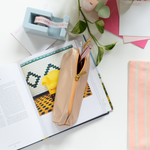 A gold metallic colored pouch with an orange zipper, displayed with metallic colored Jotter pens, a powder blue desk set piece, a book, and a plant.