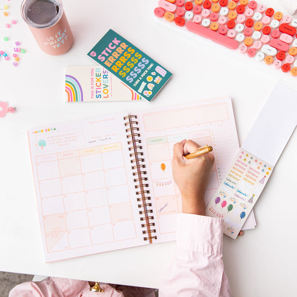 Girl writing on an undated planner with both sticker booklets and "is it friday yet?" tumbler with misc items.