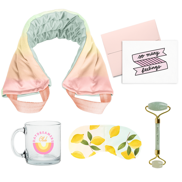 flat image of a collection of self care items including a neck wrap in a soft rainbow gradient print, a clear glass mug that says daydreamers club, a weighted eye mask with lemons imprint, a greeting card that says so many feelings, and a jade roller with roll with it imprinted in dark green. 