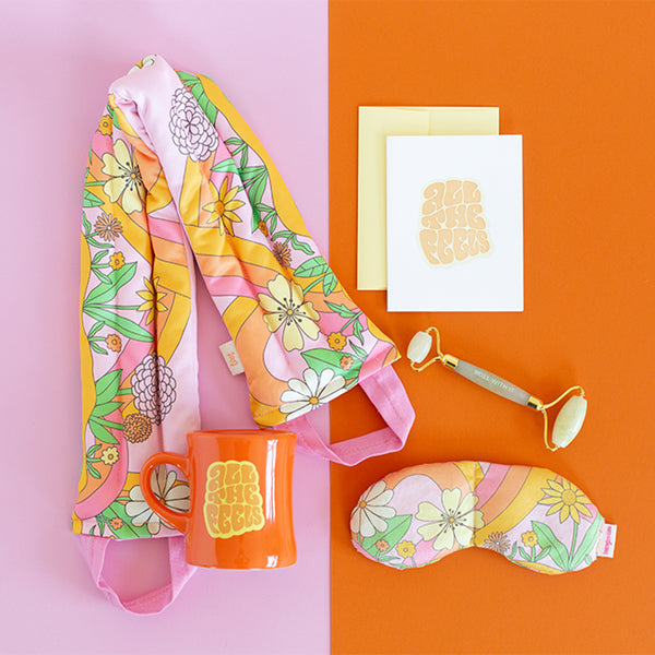 flat image of a collection of self care items including a neck wrap and an eye mask in a pink floral and swirl print, an orange diner style mug and a card that say all the feels, and a face stone roller