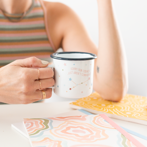 woman sitting at a table in a striped shirt holding a zodiacs not compatible campfire mug