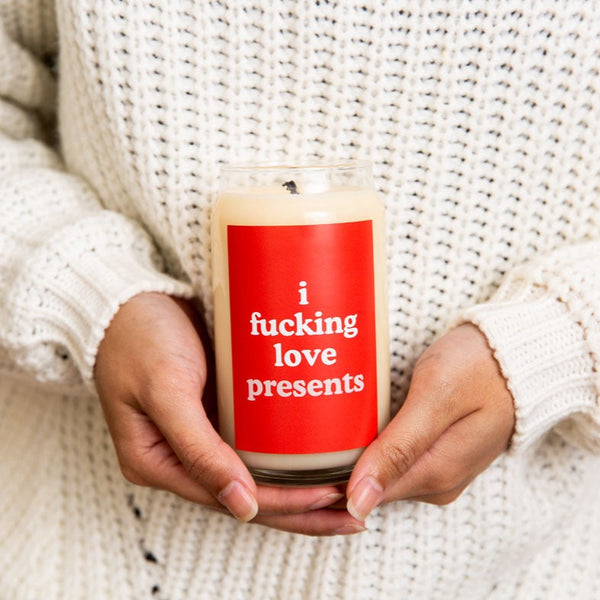 Someone holding a 12 oz. Holiday candle with a red decal that says, "i fucking love presents."