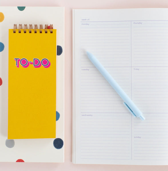 Small yellow taskpad with a magenta elastic closure and To Do written on the cover sitting on ball pit notebook